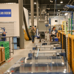 A manufacturing facility interior. TAhe space is organized, with various industrial equipment, workstations, and people engaged in different tasks. Boxes and green crates are stacked on one side, while machinery and safety barriers are positioned on the production line.