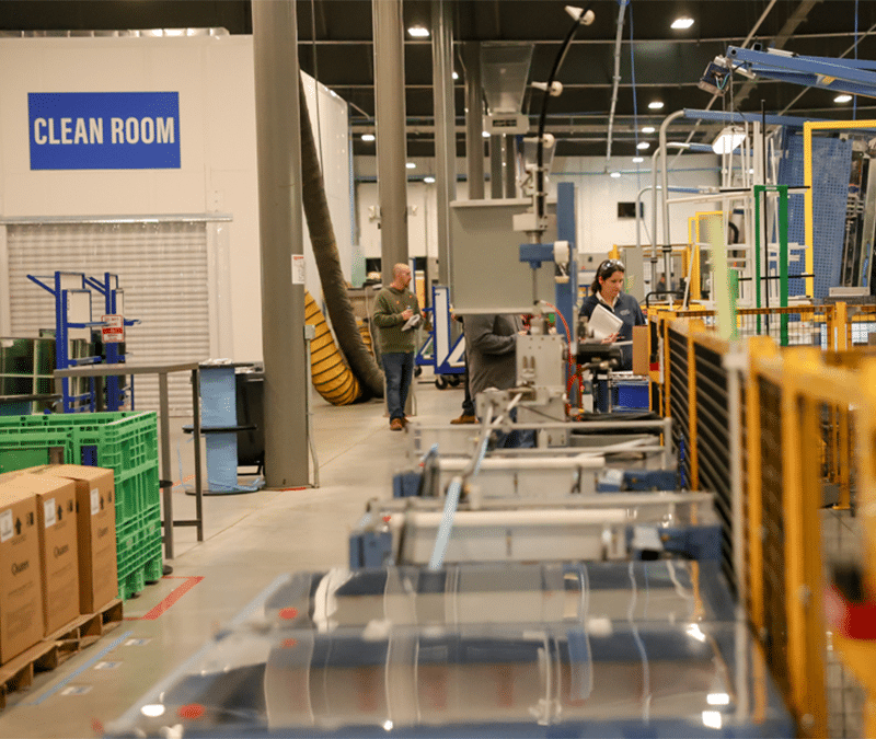 A manufacturing facility interior. TAhe space is organized, with various industrial equipment, workstations, and people engaged in different tasks. Boxes and green crates are stacked on one side, while machinery and safety barriers are positioned on the production line.