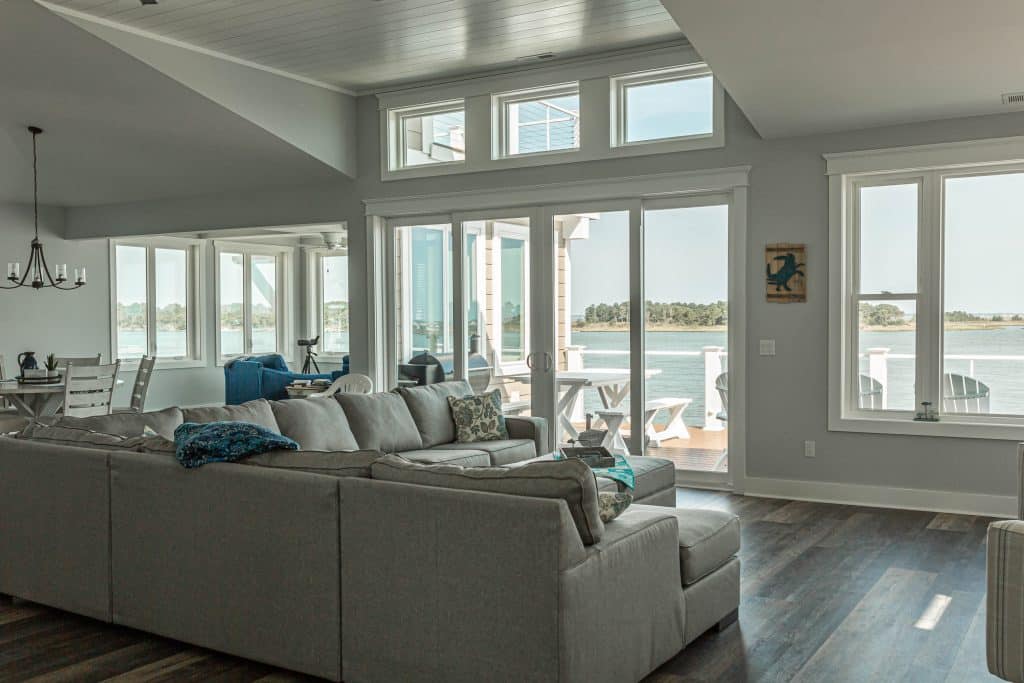 A cozy living room with a large gray sectional sofa facing expansive sliding glass doors and windows that overlook a scenic waterfront. The space has an open layout, with a dining table and chairs to the left. Light fills the room through the large windows, creating a bright and inviting atmosphere.