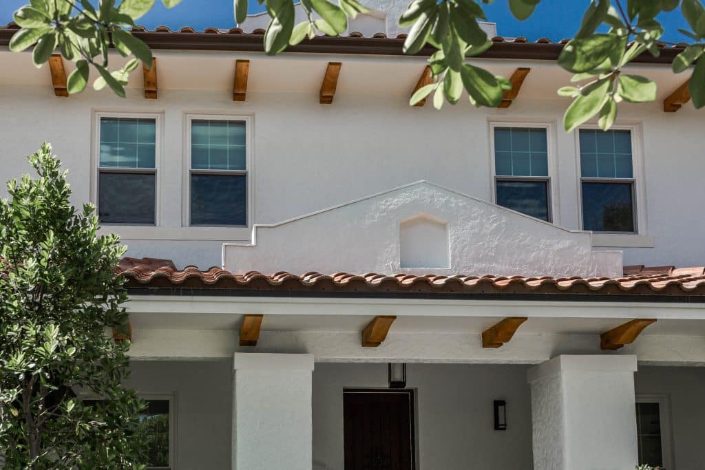 A white stucco Mediterranean-style home with two rectangular windows, exposed wooden beams, and a terracotta tiled roof. A tree partially frames the view of the house's front facade.