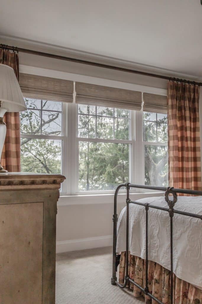 A cozy bedroom featuring large windows with an outdoor view of trees, bringing natural light into the space. The room includes an iron-framed bed with a white quilt and floral bed skirt. The windows are dressed with light-colored roman shades and checkered curtains, creating a warm and rustic ambiance.