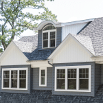A close-up of a home's upper exterior featuring a stylish, multi-gabled roof with gray shingles and white trim. The house has several large, white-framed windows, including a distinctive arched dormer window, adding architectural interest. The gray siding and detailed window design contribute to a classic and sophisticated look.