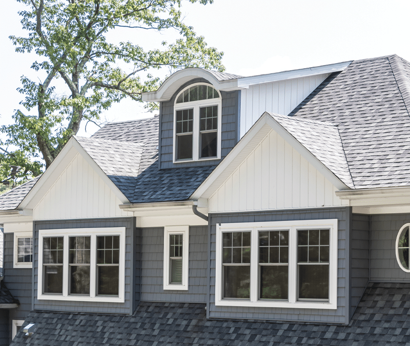 A close-up of a home's upper exterior featuring a stylish, multi-gabled roof with gray shingles and white trim. The house has several large, white-framed windows, including a distinctive arched dormer window, adding architectural interest. The gray siding and detailed window design contribute to a classic and sophisticated look.