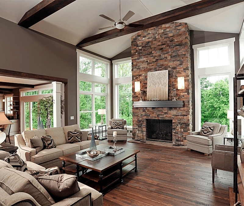 A spacious living room with vaulted ceilings features a large stone fireplace as the focal point. The room is furnished with beige sofas, patterned chairs, and a wooden coffee table, with large windows offering a view of lush greenery outside.