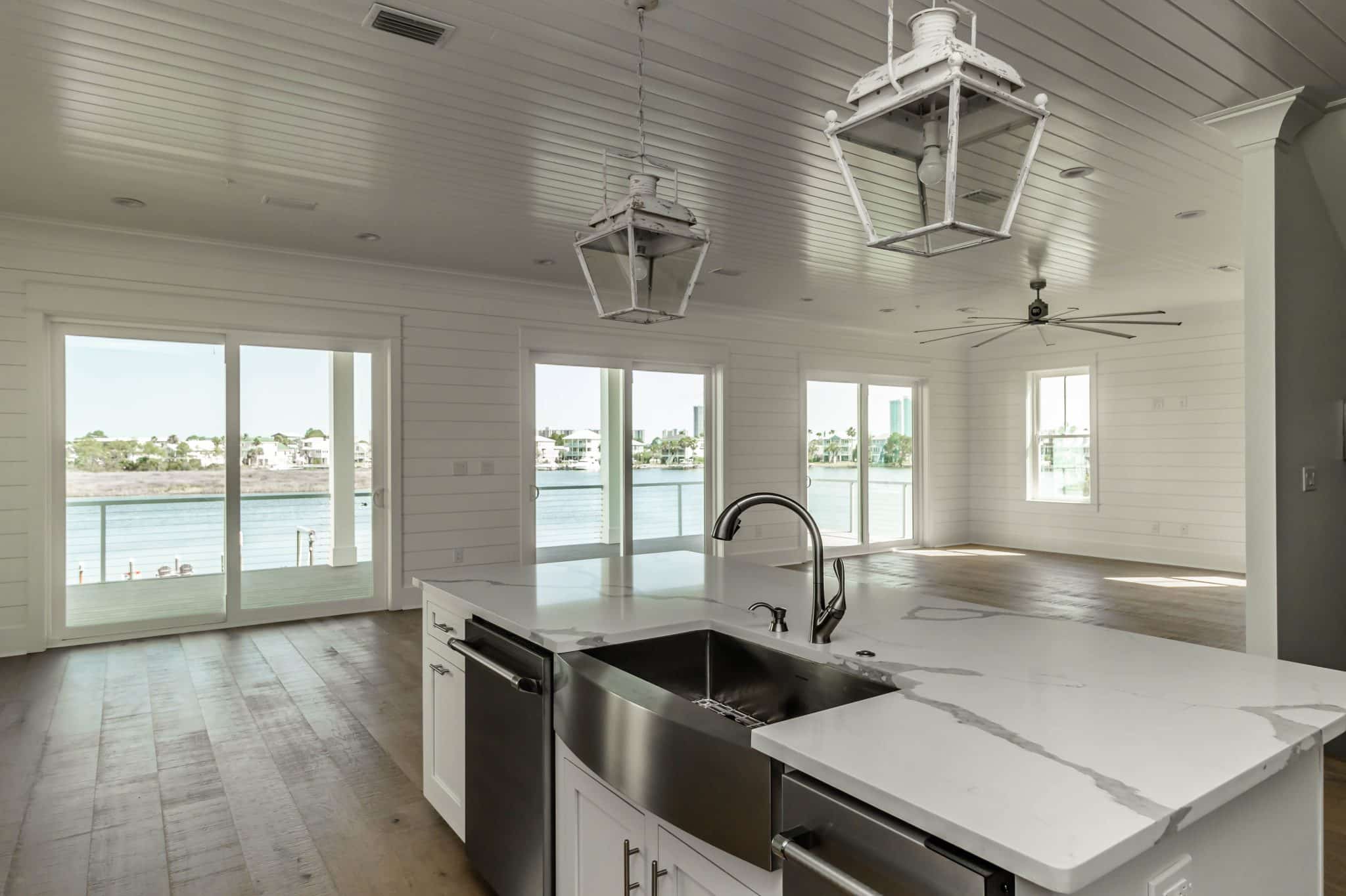 A bright, open-concept living space with large sliding glass doors offers a waterfront view. The kitchen features a marble countertop island, stainless steel sink, and modern pendant lighting.