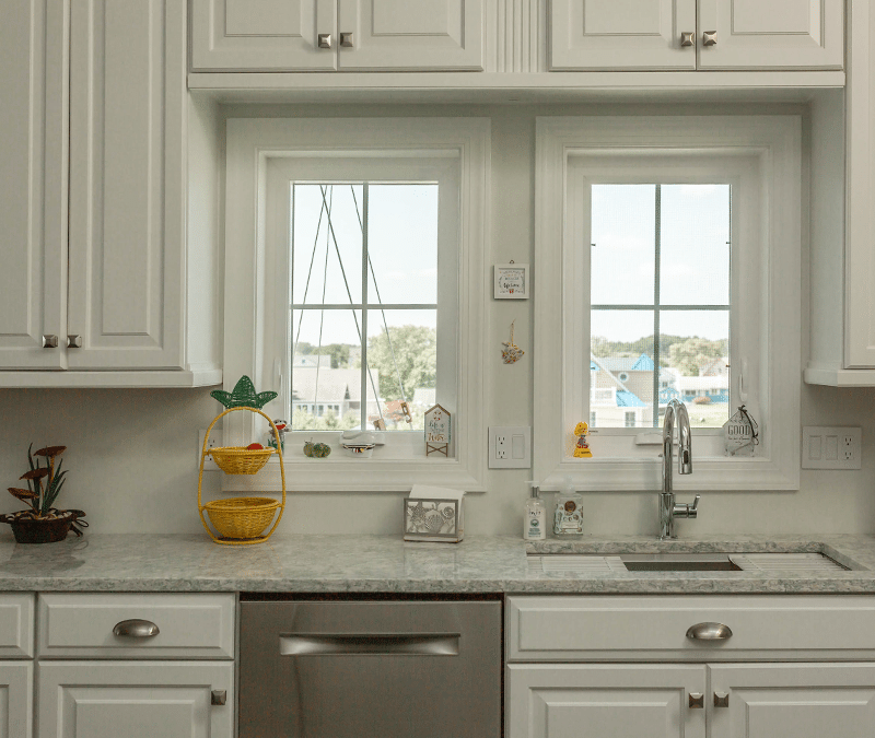 A bright kitchen with white cabinets and a marble countertop features two windows above the sink, offering a view of houses and trees outside. The countertop is decorated with a yellow basket stand and small decorative items.