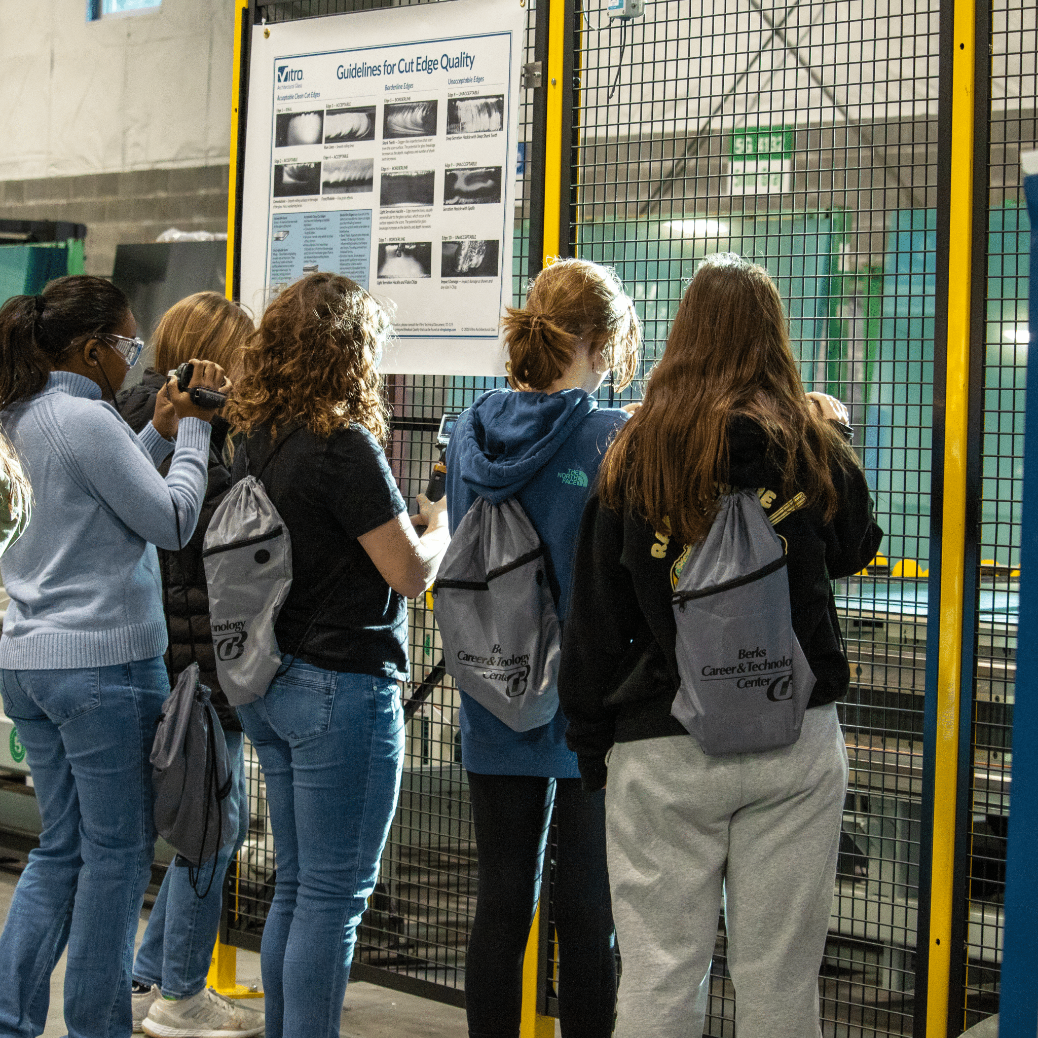 wscm_1 A group of people, wearing backpacks and safety glasses, observe machinery behind a metal safety barrier in an industrial setting. Some individuals use cameras to document the process, while a poster on "Guidelines for Cut Edge Quality" is displayed on the barrier.