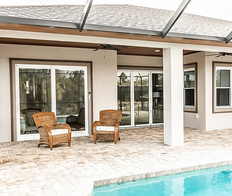 A patio area with wicker chairs overlooks a pool, featuring sliding glass doors and windows. The space is covered with a screened enclosure and brick flooring.