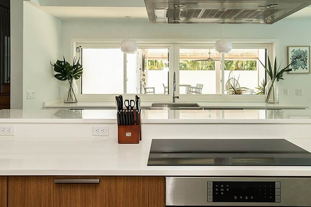 A modern kitchen with a sleek, white countertop and a stovetop in the foreground. A knife set and plants decorate the space, with large windows offering a view of an outdoor patio and greenery.