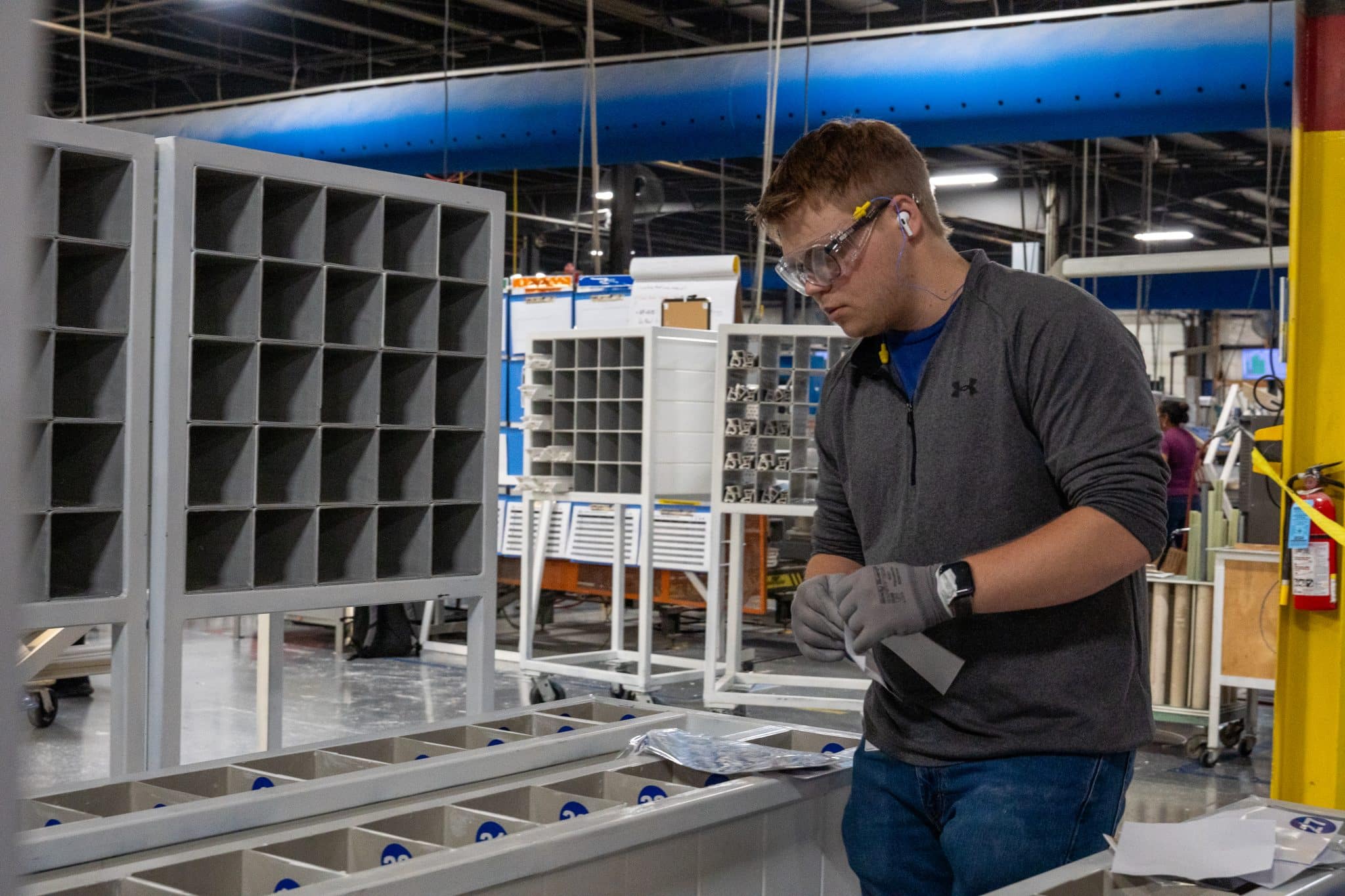 A person wearing safety goggles and gloves works in a factory, organizing materials into labeled compartments. The workspace is surrounded by shelves and industrial equipment.