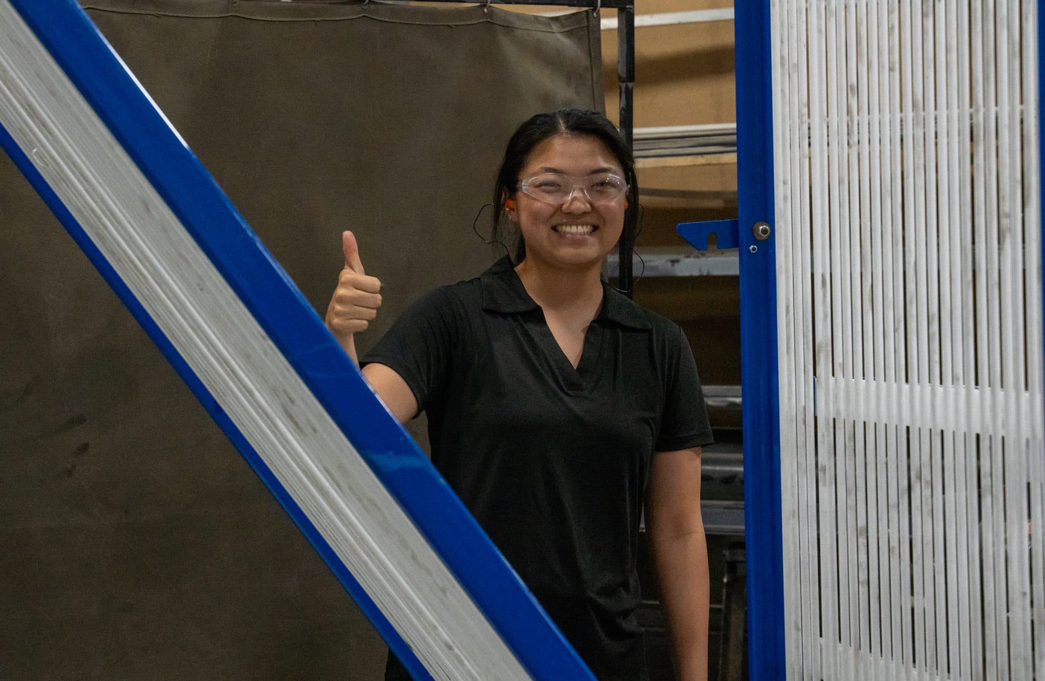 A person wearing safety goggles gives a thumbs-up while standing next to blue equipment in a workshop, smiling confidently.