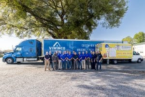 A group of people stands in front of two trucks, one branded for "Viwinco Windows + Doors" and the other for Hometown Contractors Inc, a Viwinco Blue Label Master Installer They are smiling and posing together under the shade of a large tree.