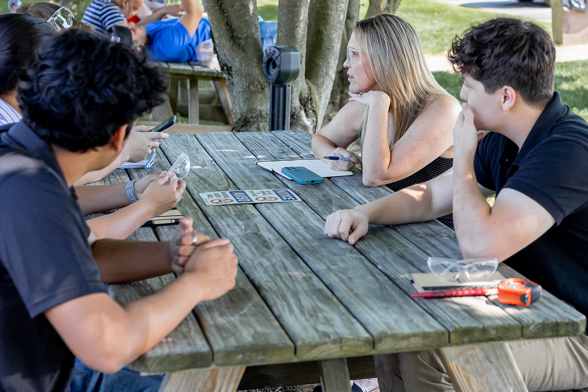 A group of people sitting at a wooden picnic table outdoors, engaged in conversation. One person holds safety goggles, while another has a pen and notebook in front of them.
