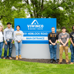 A group of six people stands in front of a blue "Viwinco Windows + Doors" sign, located at 851 Hemlock Road. They are smiling and wearing casual attire.