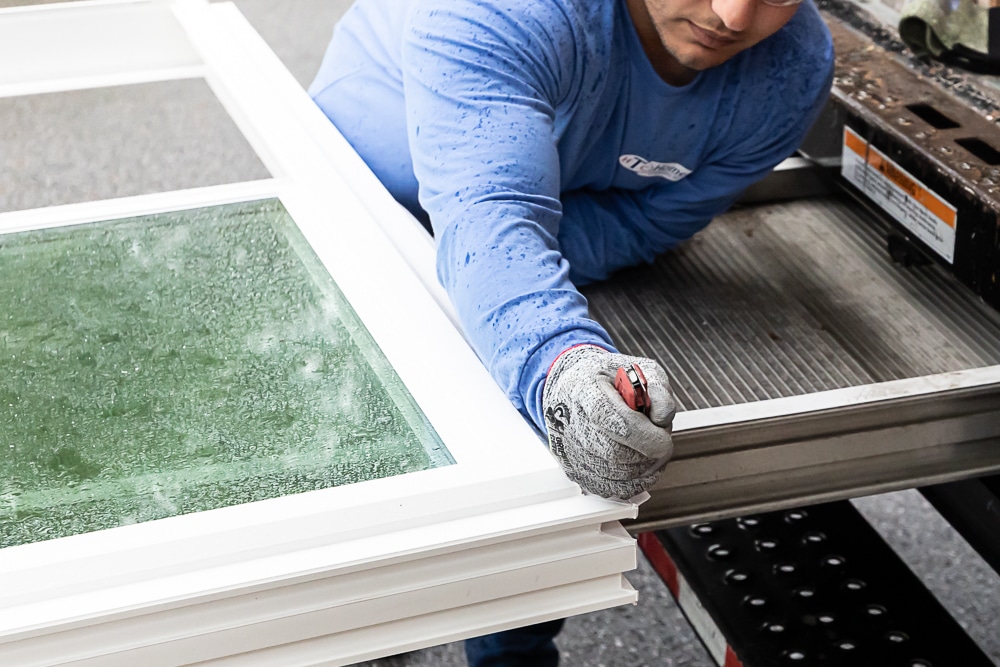 Hometown Install Day 1 - Rain Web-16 A person in a blue shirt is preparing a window frame with a damp glass panel, using a utility knife while standing next to a truck bed.