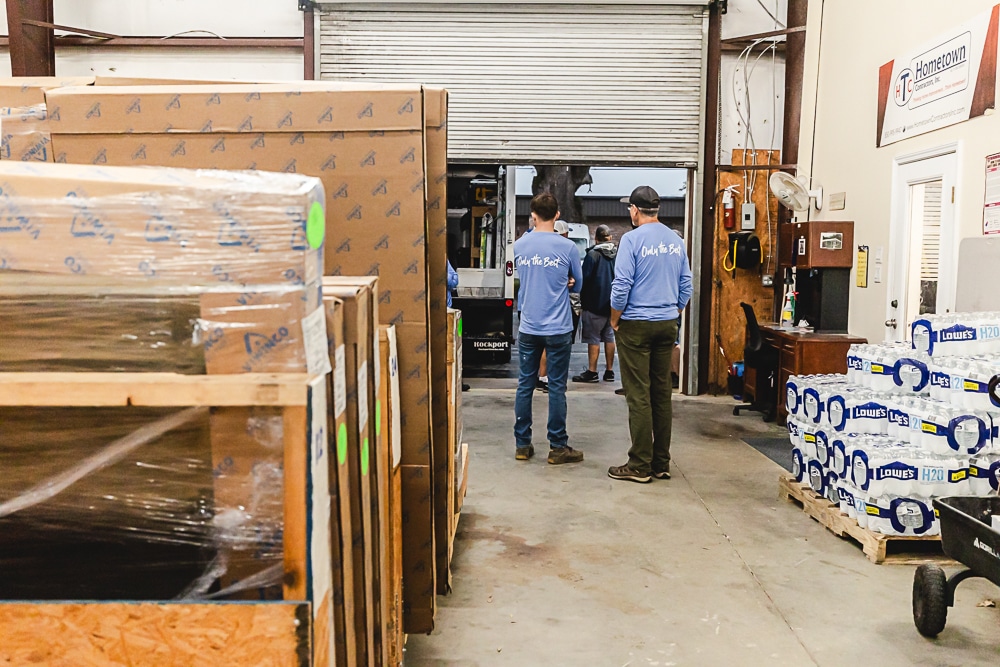 Workers in blue shirts labeled "Only the Best" stand in a warehouse near a stack of packaged goods and pallets of water bottles, with a forklift and open garage door in the background.