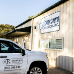 A white company truck is parked in front of a beige building with the sign "Hometown Contractors, Inc." on the side. The truck displays the company's logo and contact information.