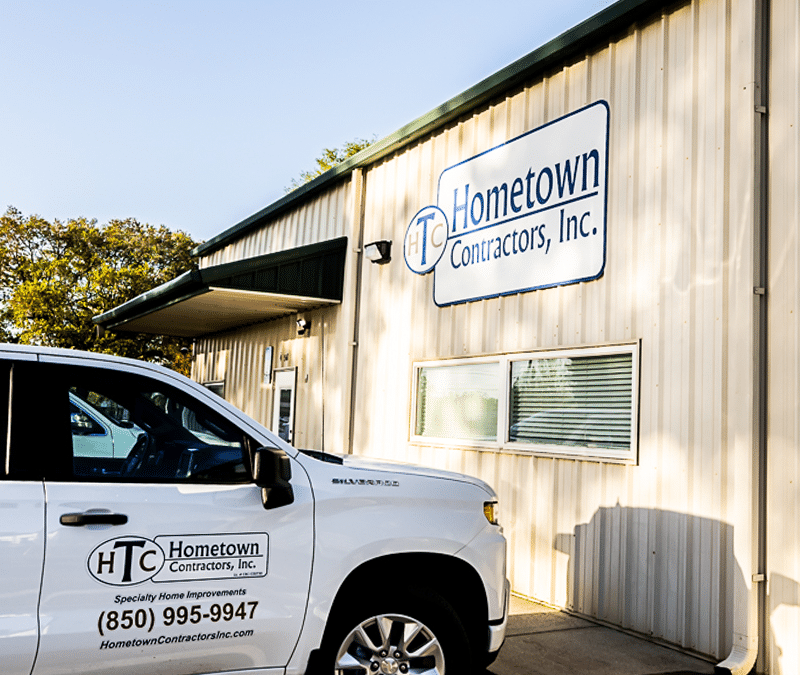 A white company truck is parked in front of a beige building with the sign "Hometown Contractors, Inc." on the side. The truck displays the company's logo and contact information.