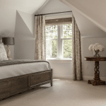A cozy bedroom with slanted ceilings, featuring a wooden bed, neutral bedding, and a small table with a floral arrangement near a window with curtains.