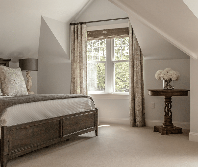 A cozy bedroom with slanted ceilings, featuring a wooden bed, neutral bedding, and a small table with a floral arrangement near a window with curtains.