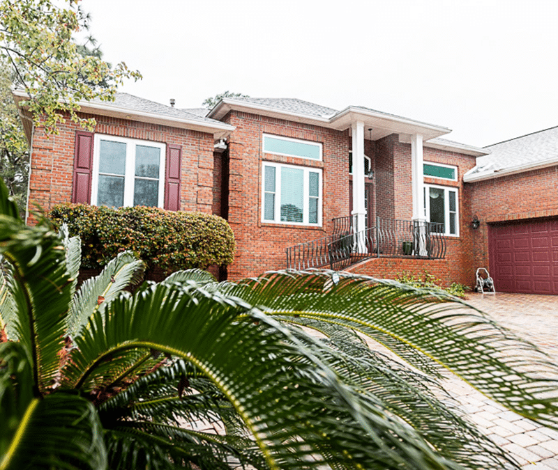A brick home with a large driveway made of stone pavers, featuring a front porch with columns and well-maintained greenery, including a large palm.