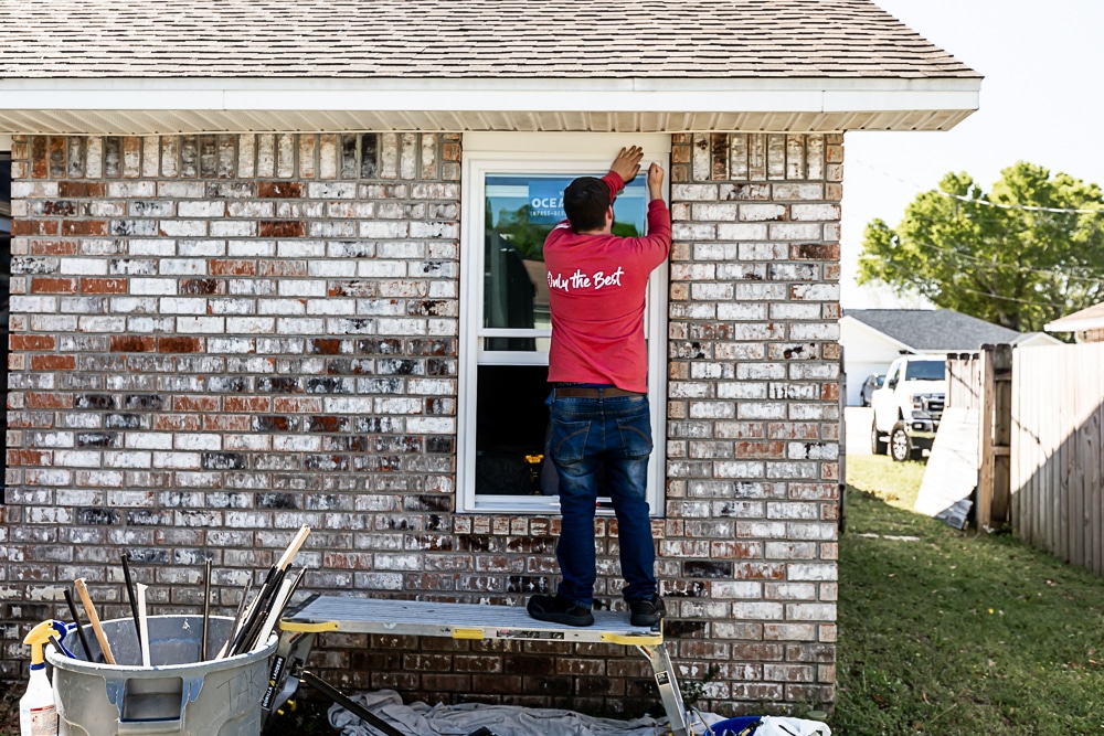 A worker in a red shirt is standing on a platform, installing or adjusting a window on a brick house. Tools are scattered in a nearby bin.