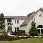A large, two-story white brick house with landscaped bushes and trees sits on a well-maintained lawn with a walkway leading to the front door.