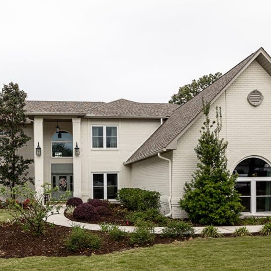 A large, two-story white brick house with landscaped bushes and trees sits on a well-maintained lawn with a walkway leading to the front door.