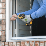 A person wearing gloves is using a power drill to install a window frame on a brick building exterior.
