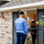 A man in a blue shirt with "Only the Best" on the back replaces the outside a window, holding a tool, while another person is reflected inside the house.