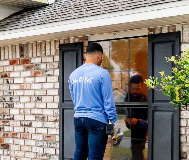 A man in a blue shirt with "Only the Best" on the back replaces the outside a window, holding a tool, while another person is reflected inside the house.